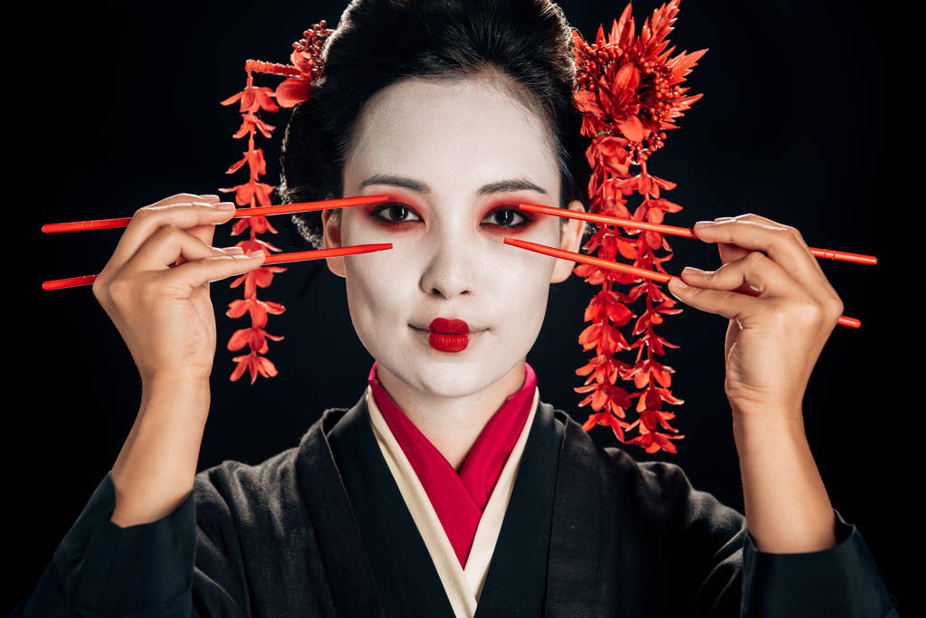 beautiful geisha in black kimono with red flowers in hair holding chopsticks near eyes isolated on black - Photo, Image