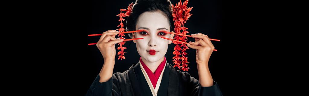 belle geisha en kimono noir avec des fleurs rouges dans les cheveux tenant baguettes près des yeux isolés sur noir, vue panoramique
 - Photo, image