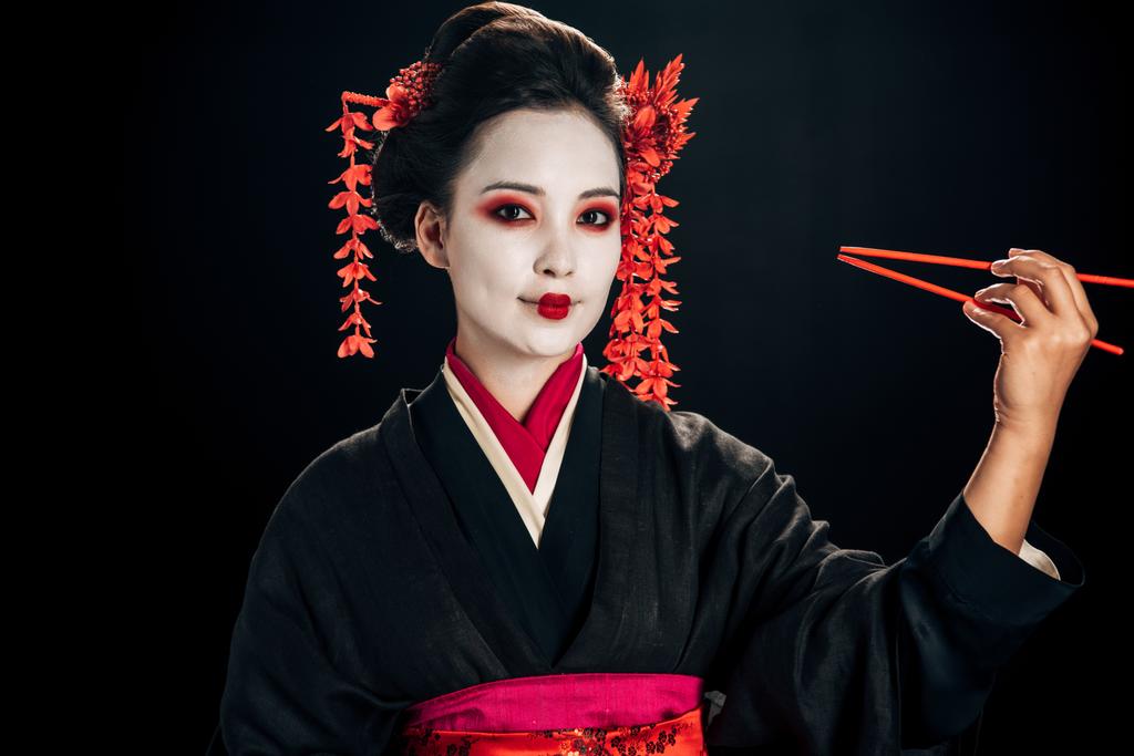 veselá a krásná gejša v černém kimonu s červenými květy ve vlasech s hůlky, izolované na černém - Fotografie, Obrázek