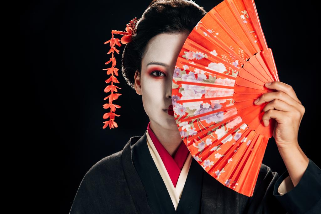 attractive geisha in black kimono with flowers in hair holding bright hand fan isolated on black - Photo, Image