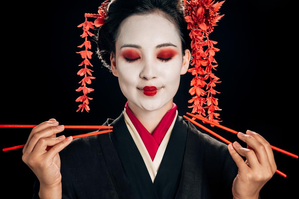 uśmiechnięta piękna gejsza w czarnym kimono z czerwonymi kwiatami we włosach i zamkniętymi oczami trzymającą pałeczki na czarno - Zdjęcie, obraz