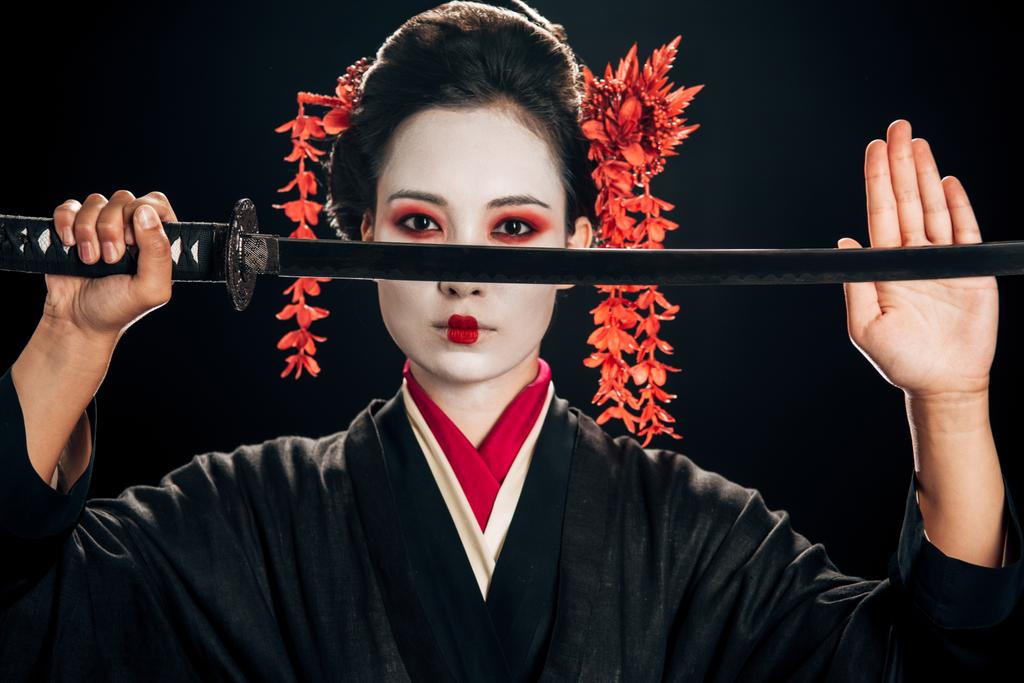 selbstbewusste Geisha im schwarzen Kimono mit roten Blumen im Haar, die Katana isoliert auf Schwarz hält - Foto, Bild