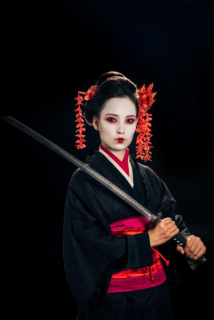 beautiful geisha in black kimono with red flowers in hair holding katana isolated on black - Photo, Image