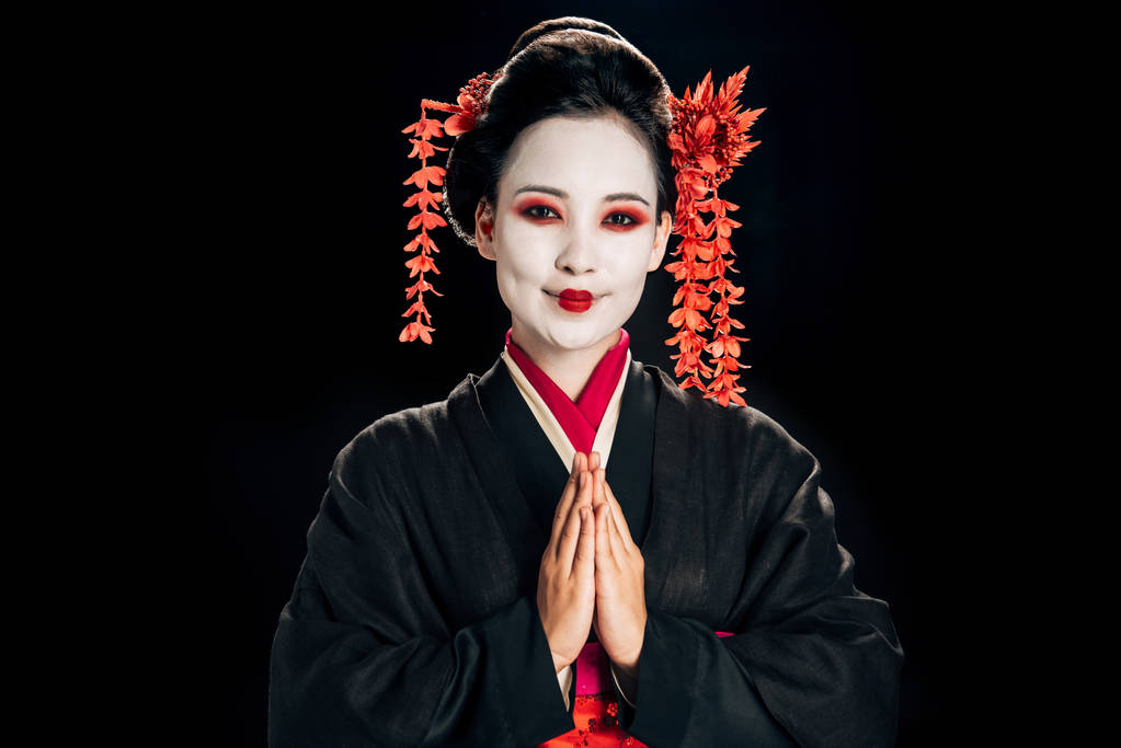 vista lateral da bela gueixa feliz em quimono preto e vermelho e flores no cabelo olhando para a câmera e saudação isolada no preto
 - Foto, Imagem