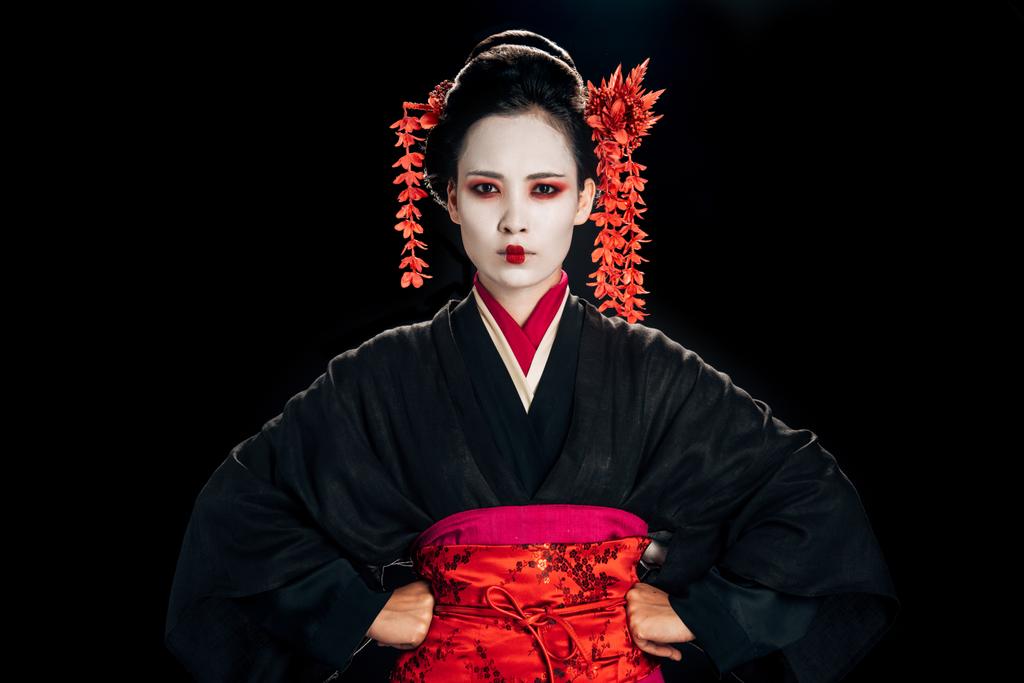 geisha hermosa enojada en kimono negro y rojo y flores en el pelo con las manos en las caderas aisladas en negro
 - Foto, Imagen