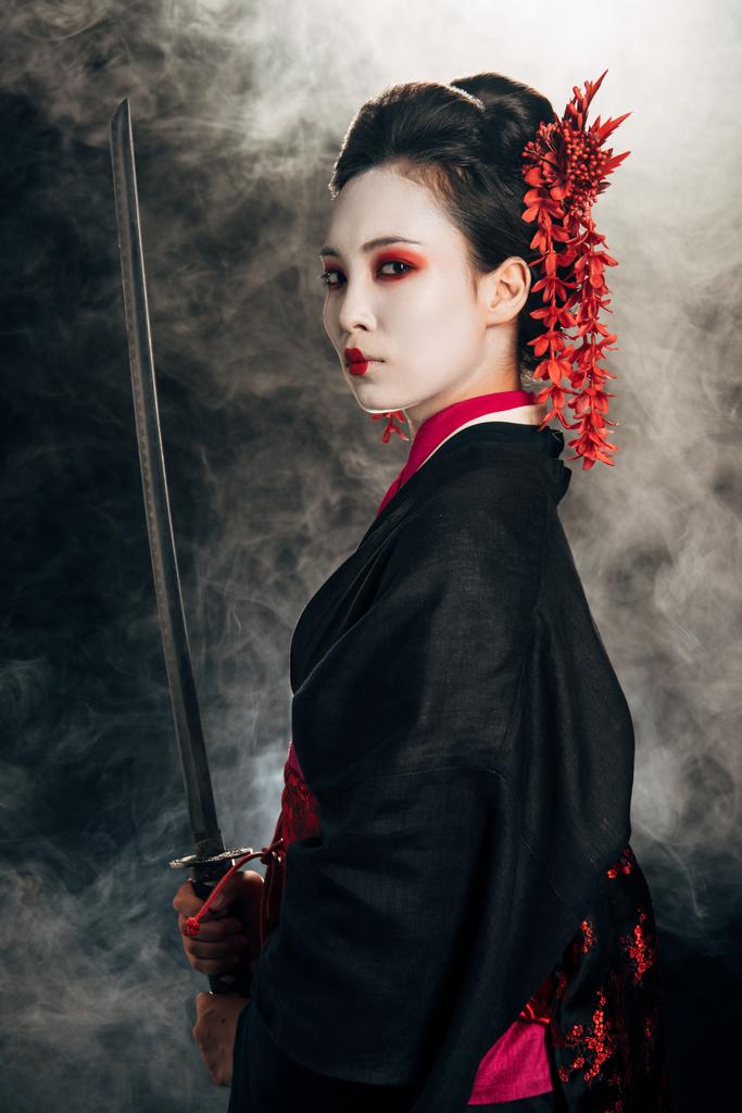 vue latérale de la belle geisha en kimono noir tenant katana en fumée sur fond noir
 - Photo, image