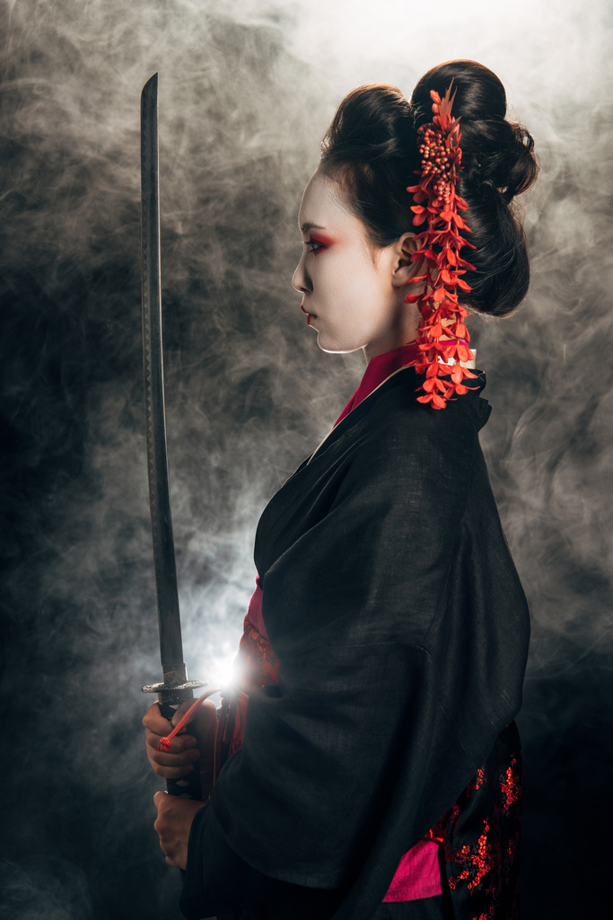 Zijaanzicht van Geisha in zwarte kimono Holding Katana in rook op zwarte achtergrond - Foto, afbeelding
