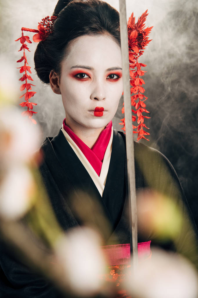 selective focus of confident geisha in black kimono holding katana in smoke and sakura branches - Photo, Image