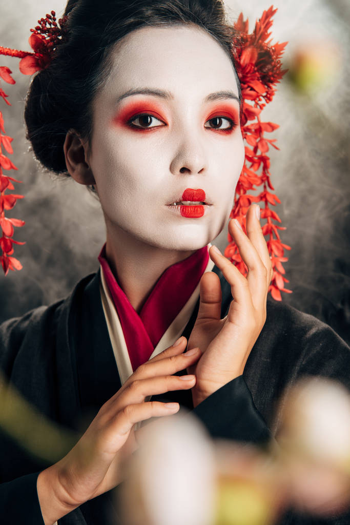 selective focus of beautiful geisha in black kimono with red flowers in hair touching face and sakura branches on black background with smoke - Photo, Image