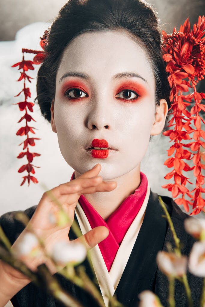 portrait of beautiful geisha in black kimono with red flowers in hair and sakura branches on black background with smoke - Photo, Image