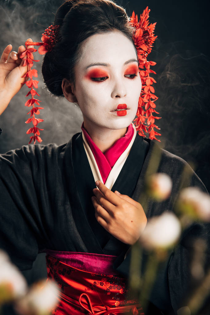 selective focus of geisha in black kimono with red flowers in hair and sakura branches on black background with smoke - Photo, Image