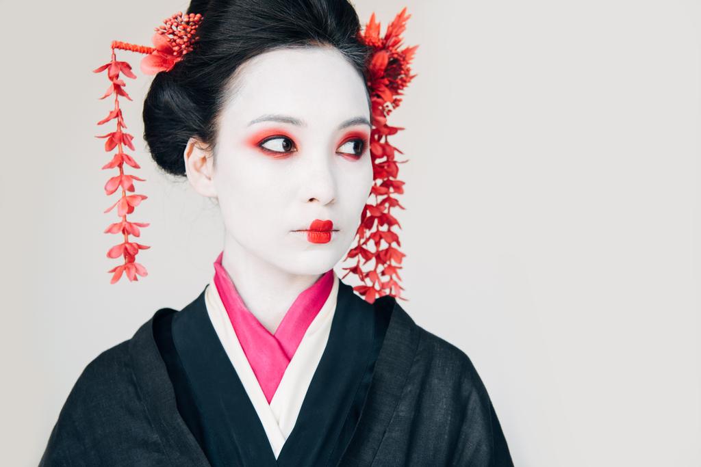 schöne Geisha im schwarzen Kimono mit roten Blüten im Haar, die isoliert auf weißem Grund wegschauen - Foto, Bild
