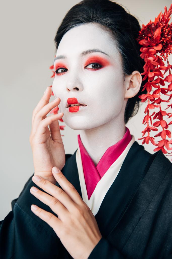 όμορφη γκέισα σε μαύρο κιμονό με κόκκινα λουλούδια στα μαλλιά αγγίζοντας το πρόσωπο απομονωθεί σε λευκό - Φωτογραφία, εικόνα