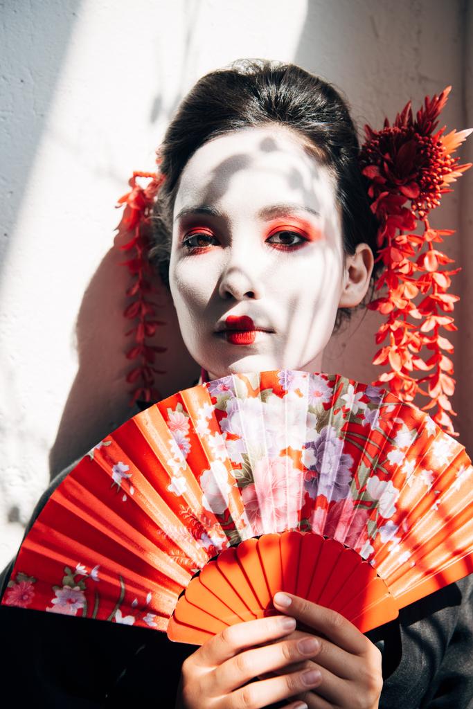 портрет красивой гейши с красным и белым макияжем, держащей за руку веер на солнце
 - Фото, изображение