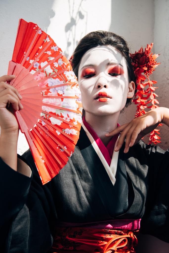 portrait of beautiful geisha with red and white makeup holding hand fan and gesturing in sunlight - Photo, Image