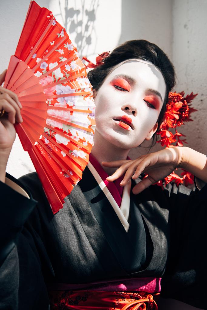 Porträt einer wunderschönen Geisha mit rot-weißem Make-up, die einen Fächer an der Hand hält und das Gesicht im Sonnenlicht berührt - Foto, Bild