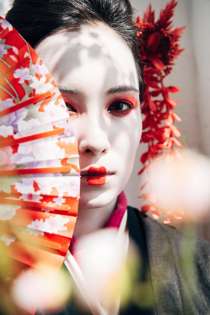 selective focus of sakura and beautiful geisha with red and white makeup holding hand fan in sunlight - Photo, Image