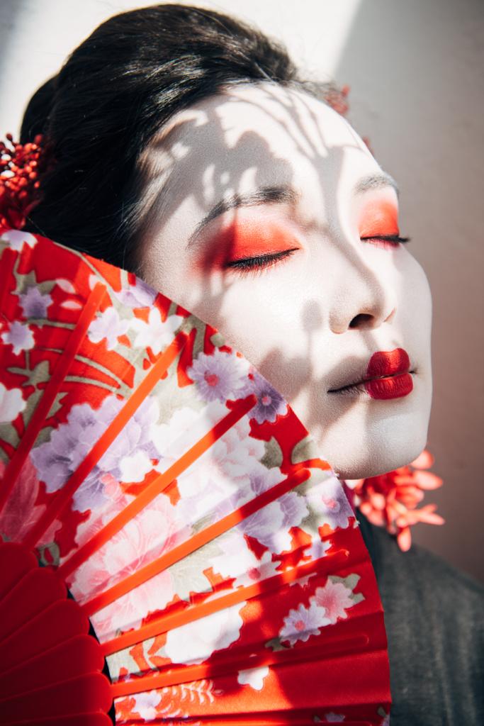 πορτρέτο της όμορφης γκέισα με κόκκινο και λευκό μακιγιάζ και κλειστά μάτια κρατώντας ανεμιστήρα χεριού στο ηλιακό φως - Φωτογραφία, εικόνα