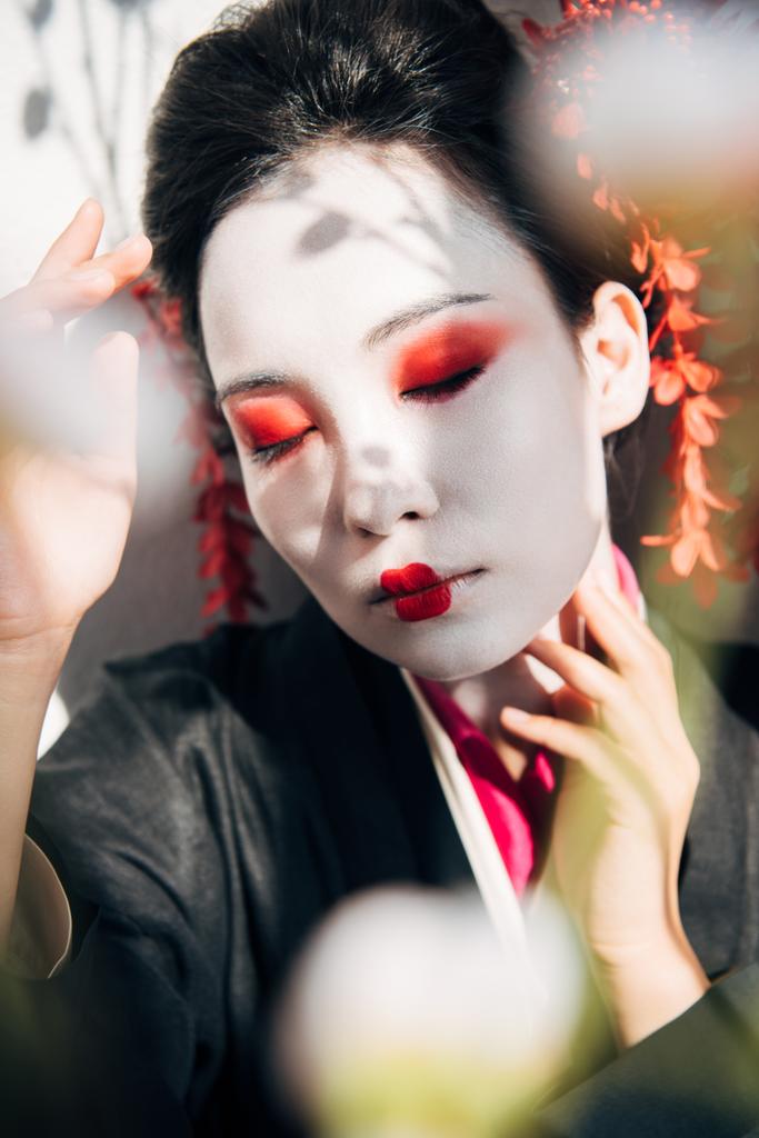 foco seletivo de galhos de árvores e bela gueixa com maquiagem vermelha e branca à luz solar
 - Foto, Imagem
