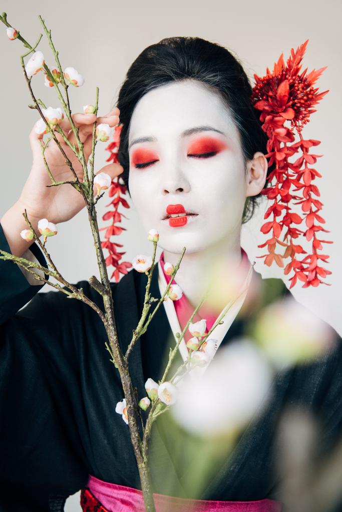 selective focus of tree branches and beautiful geisha with red and white makeup and closed eyes isolated on white - Photo, Image