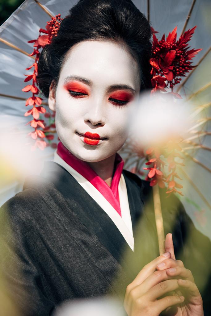 selectieve focus van boomtakken en mooie glimlachende Geisha met paraplu en gesloten ogen in zonlicht - Foto, afbeelding