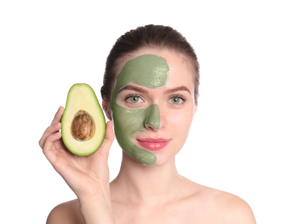 Молода жінка з глиняною маскою на обличчі тримає авокадо на білому тлі. Догляд за шкірою
 - Фото, зображення