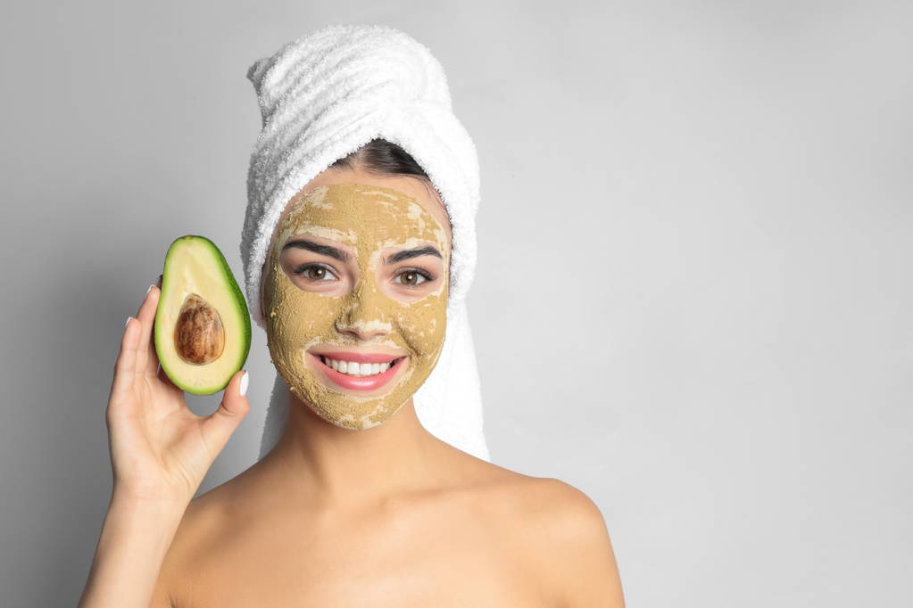 Young woman with clay mask on her face holding avocado against light background, space for text. Skin care - Photo, Image