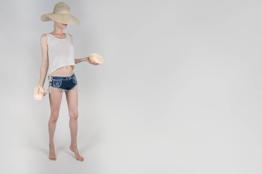 das Mädchen in Shorts und Hut, die ihr Gesicht verdecken und die Muscheln auf weißem Hintergrund isoliert halten - Foto, Bild