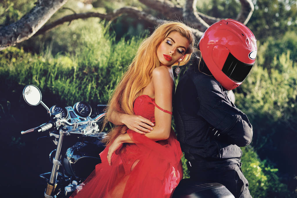 Lady wearing red dress sitting on motorcycle with man, girl leaned on guy hides his face behind helmet pose on nature, sunny summer day two people outdoors near bike, hobby lifestyle fashion concept - Photo, Image