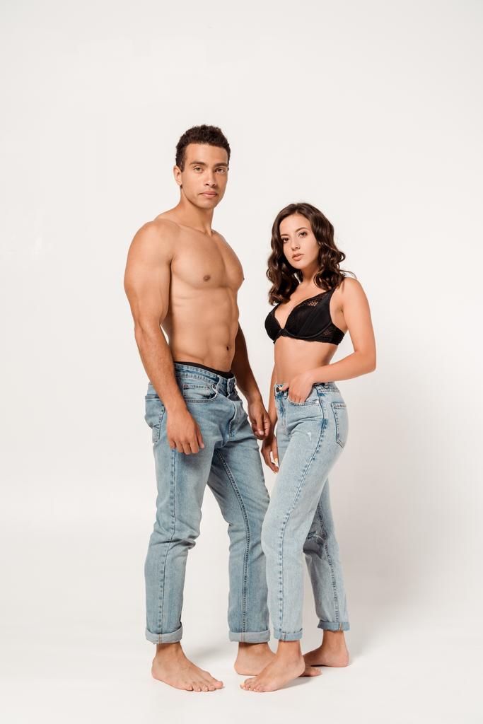 sexy woman in lace black bra and jeans standing near muscular mixed race man on white  - Photo, Image