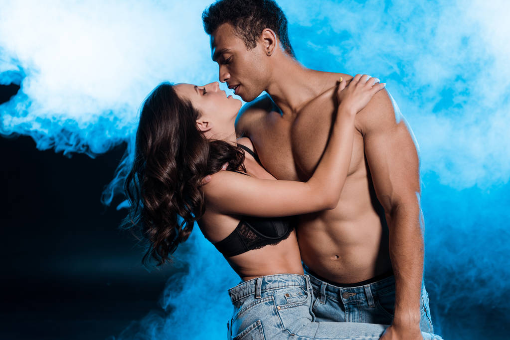 knappe gemengde race man knuffelen mooi en sexy meisje in beha op blauw met rook  - Foto, afbeelding