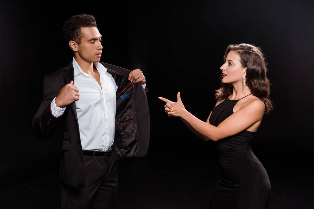 приваблива дівчина вказує пальцем на змішану расу чоловік торкається костюма ізольовано на чорному
  - Фото, зображення