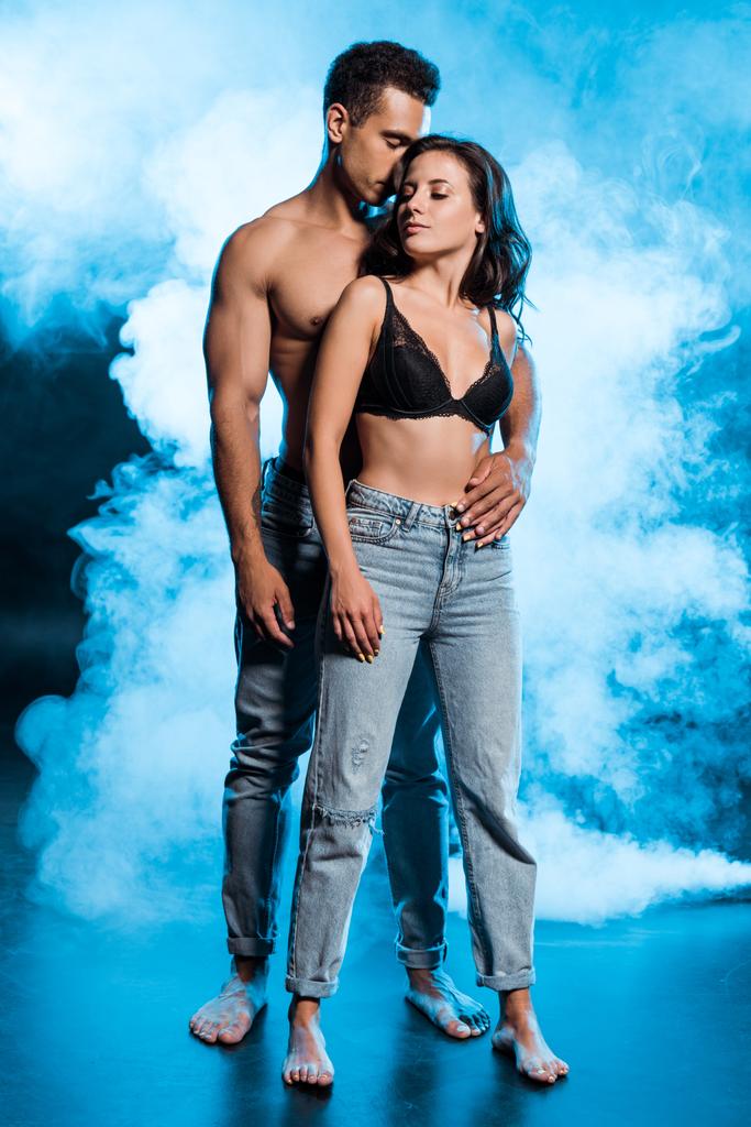sin camisa mezclado raza hombre abrazando sexy chica en denim jeans de pie en azul con humo
  - Foto, Imagen
