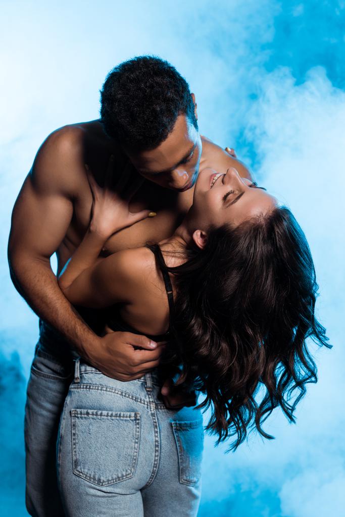 shirtless gemengde race man knuffelen gelukkig meisje in denim jeans staande op blauw met rook  - Foto, afbeelding