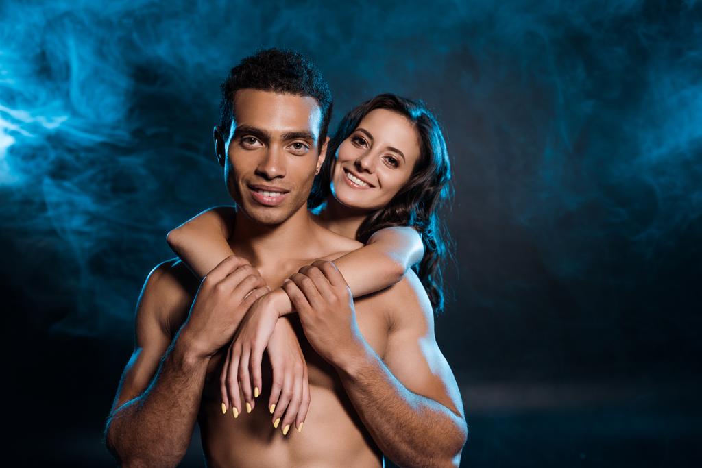 young cheerful woman hugging muscular mixed race man on black with blue smoke  - Photo, Image