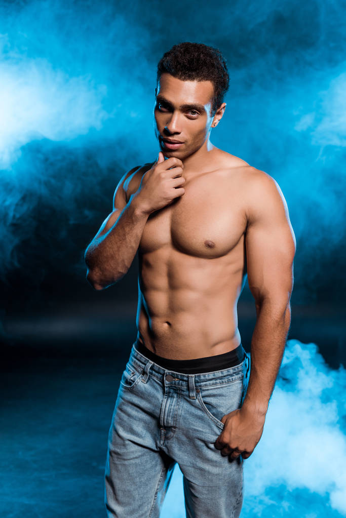 sexy Mixed Race Mann steht in Jeans und berührt Gesicht auf blau mit Rauch  - Foto, Bild