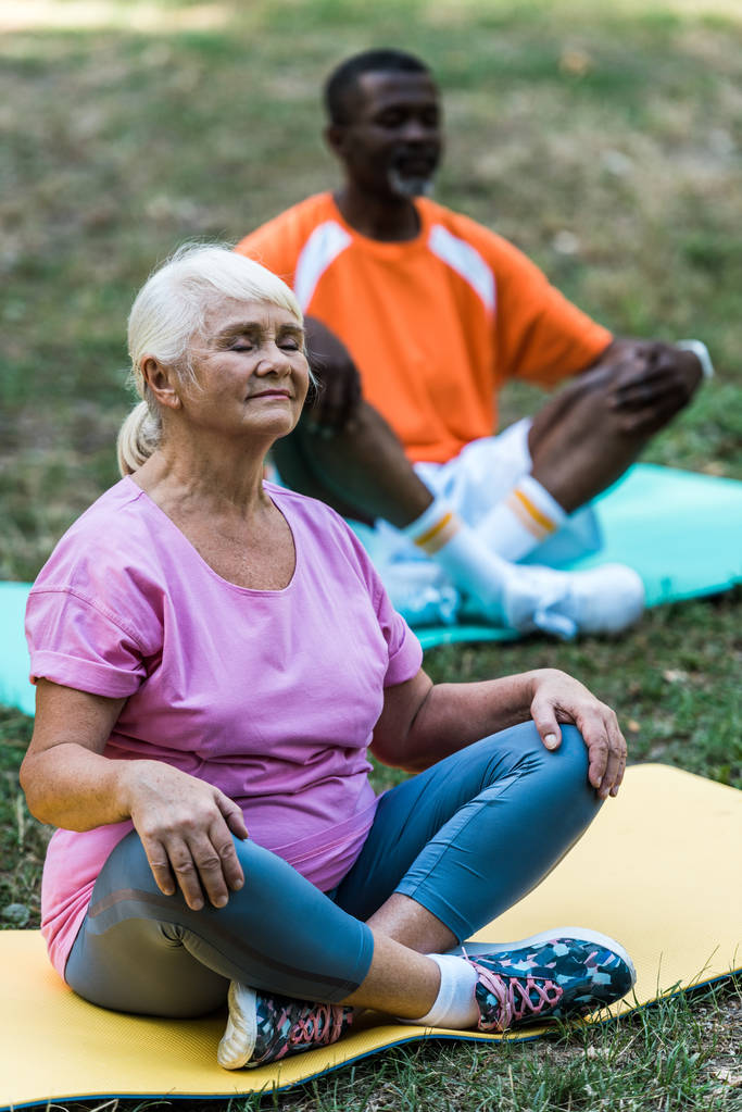 enfoque selectivo de la mujer jubilada con los ojos cerrados sentado con las piernas cruzadas cerca del hombre afroamericano senior en el parque
  - Foto, Imagen