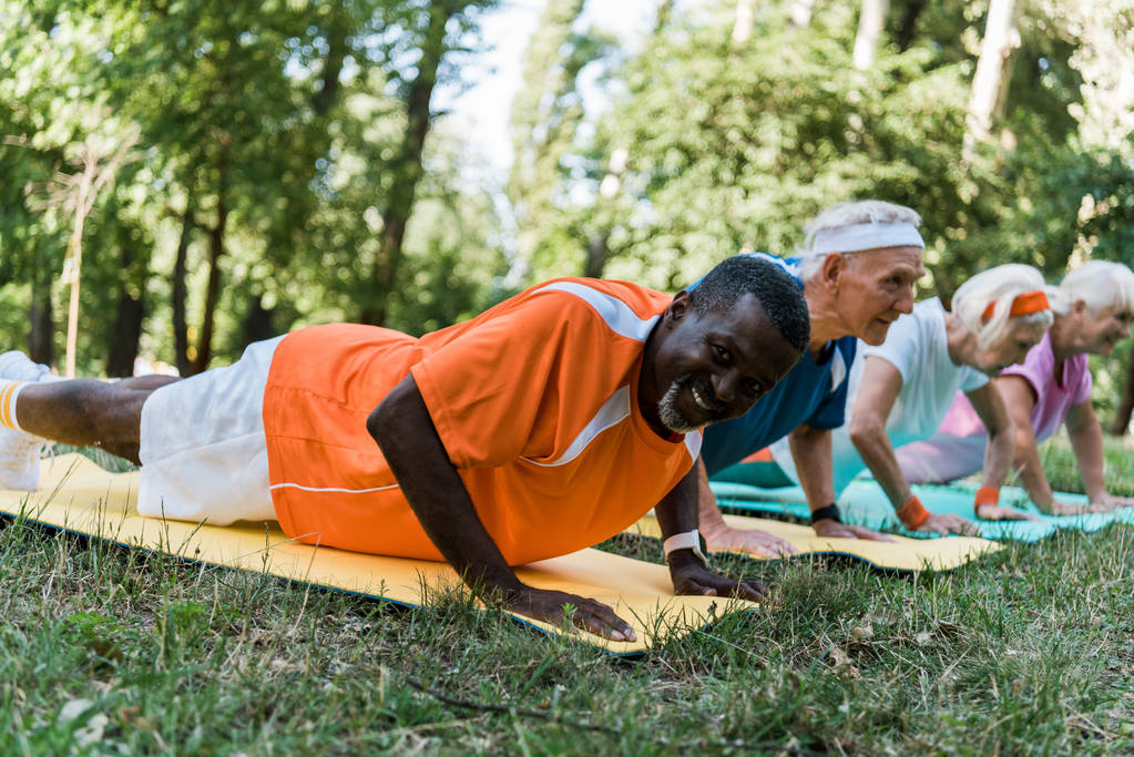 επιλεκτική εστίαση του Happy Αφρικανός Αμερικανός άνθρωπος άσκηση κοντά σε συνταξιούχοι συνταξιούχους σε χαλάκια Fitness  - Φωτογραφία, εικόνα