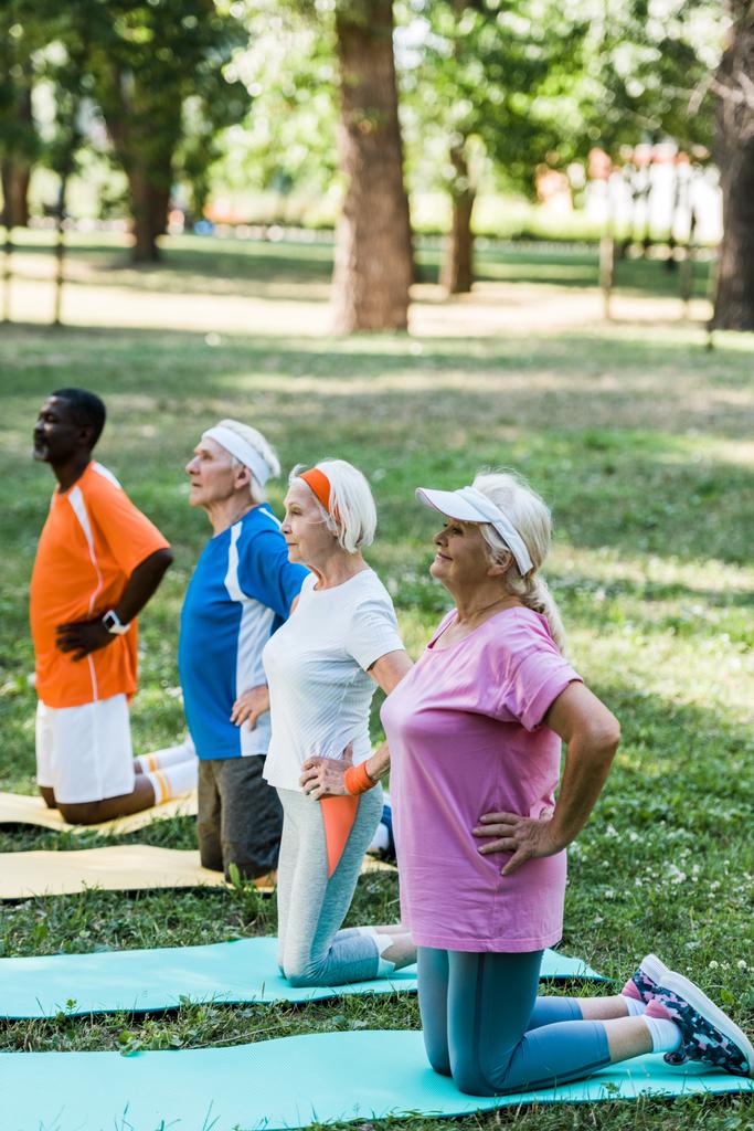 enfoque selectivo de los jubilados multiculturales felices sentados con las manos en las caderas mientras se ejercita en alfombras de fitness
  - Foto, Imagen