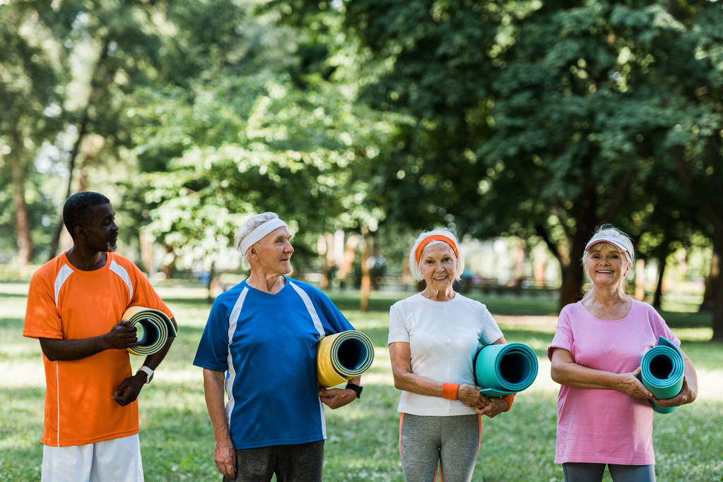 happy retired and multicultural pensioners holding fitness mats in park  - Photo, Image