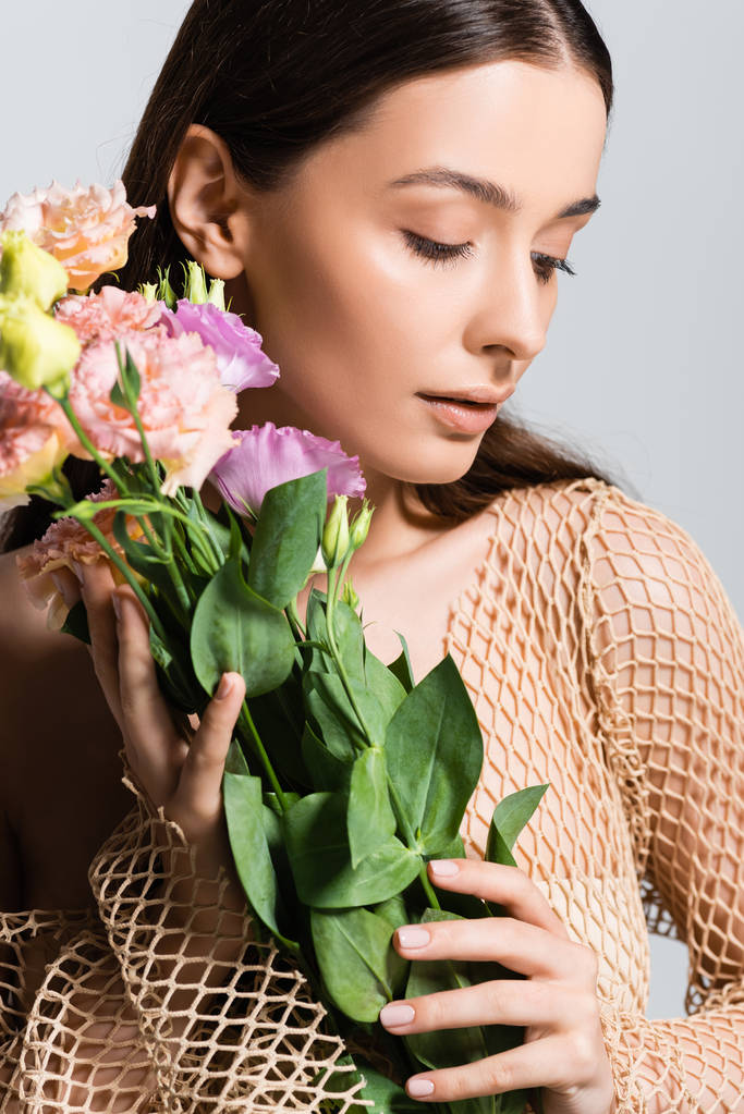 エウストマの花の花束を持ち、灰色で孤立した見下ろすベージュメッシュの美しい若い女性 - 写真・画像