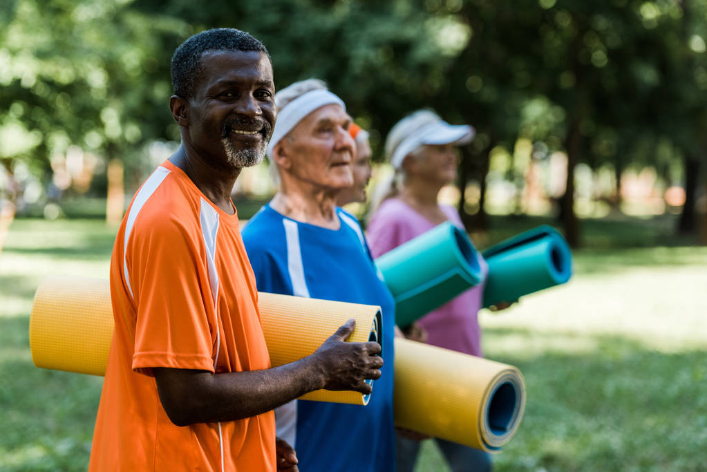 selective focus of positive retired african american man near pensioners holding fitness mats  - Photo, Image