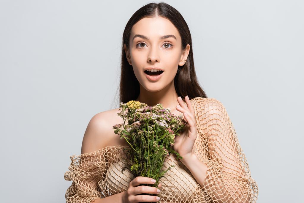 beautiful shocked woman in beige mesh with wildflowers looking away isolated on grey - Photo, Image