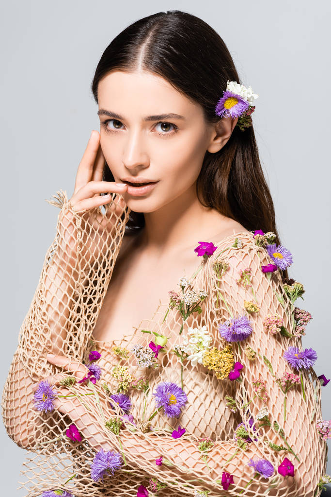 όμορφη γυναίκα σε δίχτυ μπεζ ρούχα με μωβ λουλούδια αγγίζοντας τα χείλη απομονώνονται σε γκρι - Φωτογραφία, εικόνα