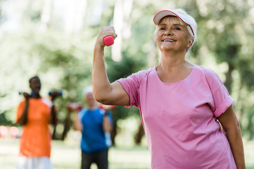 年金受給者の近くで運動しながらダンベルを保持するキャップで幸せな引退した女性の選択的な焦点  - 写真・画像