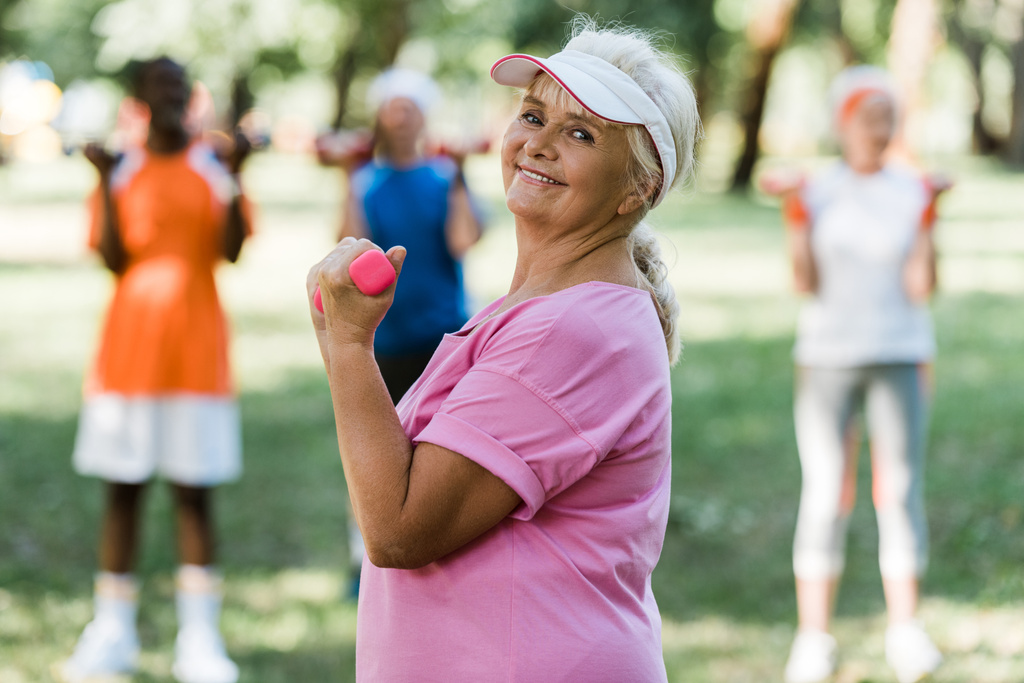 enfoque selectivo de la alegre mujer jubilada con gorra sosteniendo pesas mientras hace ejercicio cerca de los pensionistas
  - Foto, imagen