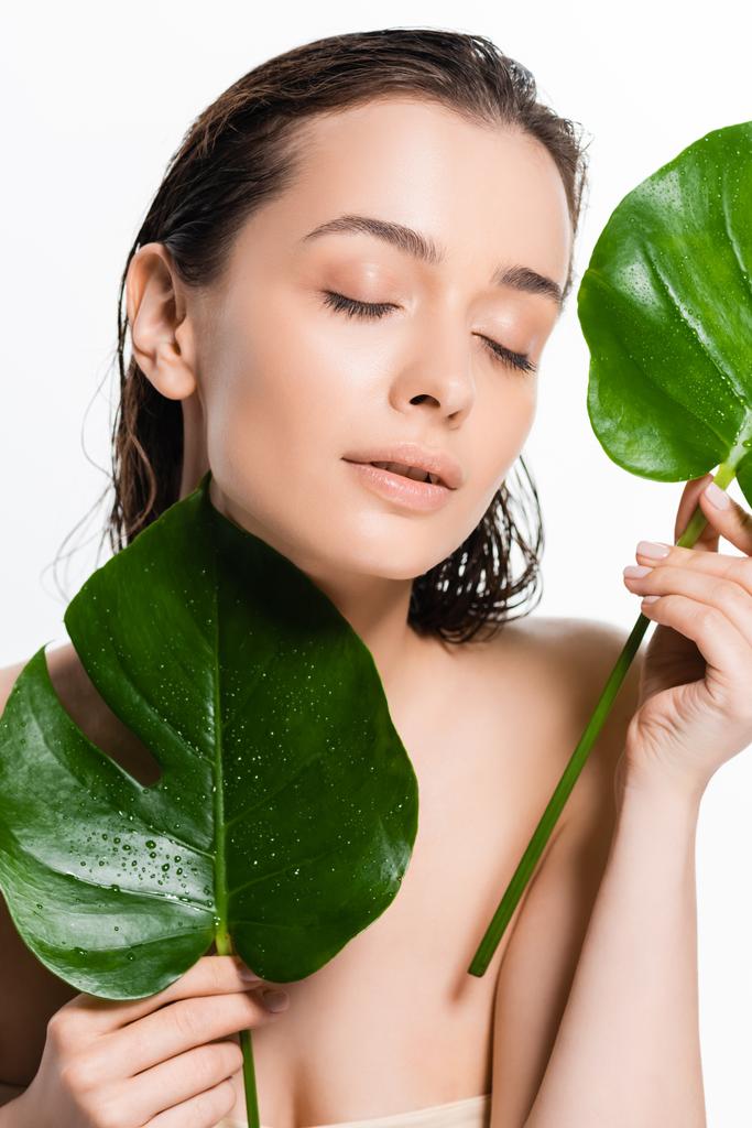 mujer joven desnuda mojada con los ojos cerrados sosteniendo hojas de palma verde con gotas de agua aisladas en blanco
 - Foto, Imagen