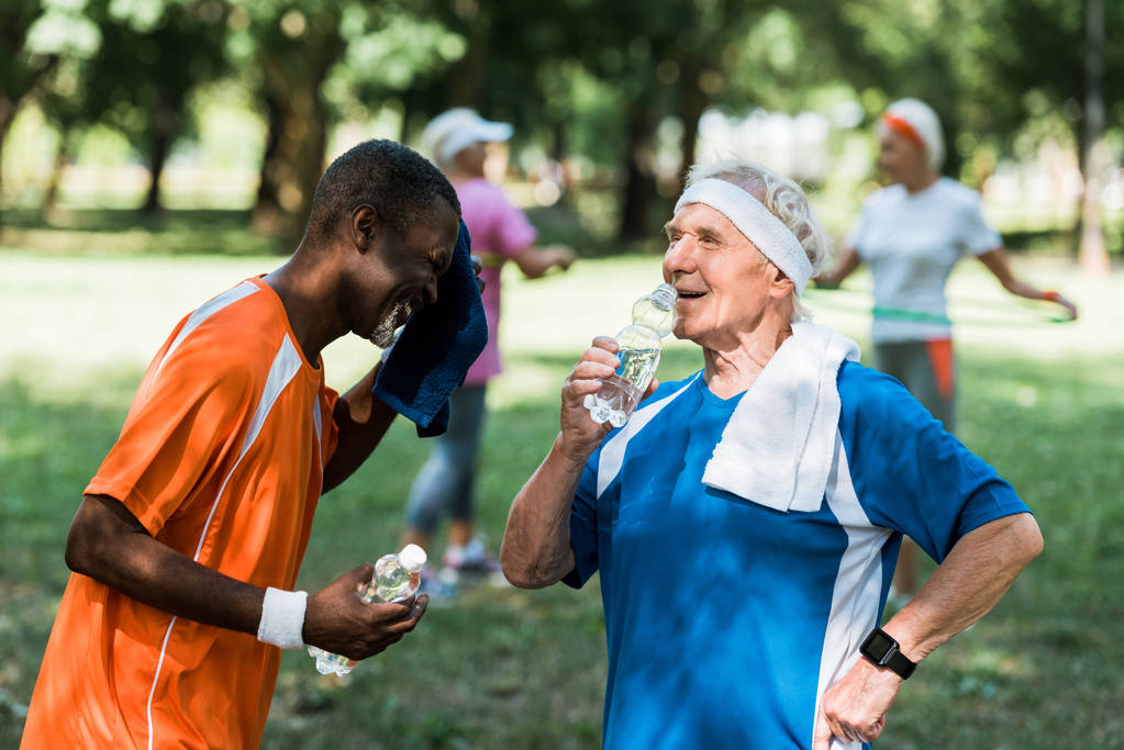 selective focus of sportive multicultural retired men holding bottles with water near senior women  - Photo, Image