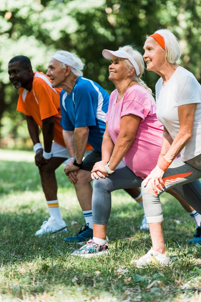 enfoque selectivo de personas mayores alegres y multiculturales haciendo ejercicio de estiramiento en la hierba
  - Foto, imagen