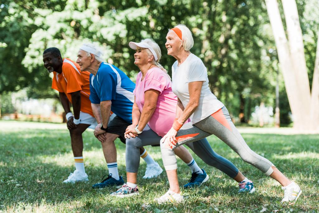 selective focus of happy retired and multicultural people doing stretching exercise on grass  - Photo, Image
